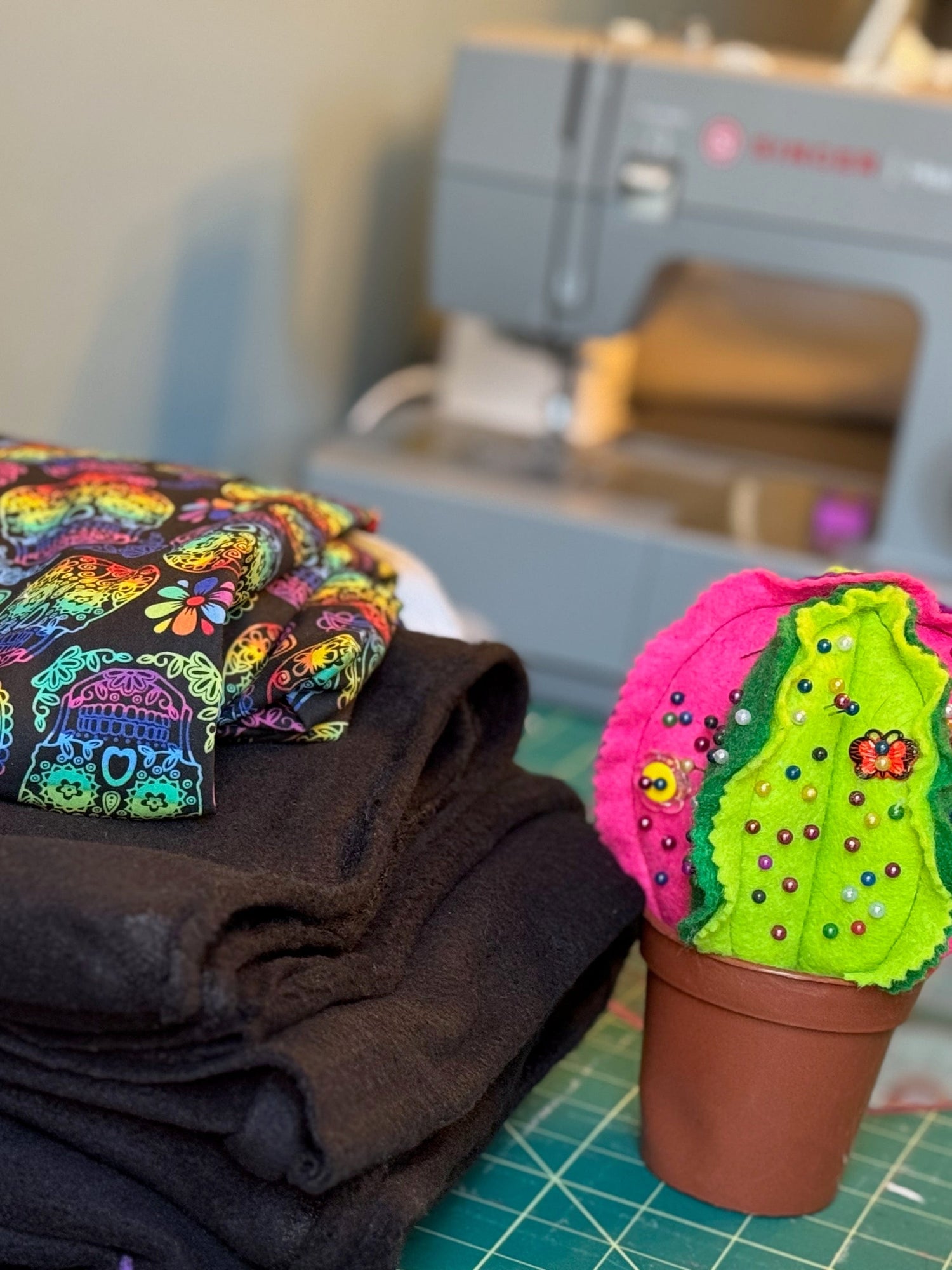 A bewitching stack of midnight-black fabric and vibrant, spellbinding prints rests beside a whimsically spooky felt cactus perched in a tiny gothic pot. In the misty background, an enchanting sewing machine lurks—ready to conjure magical accessories for your beloved small familiars.