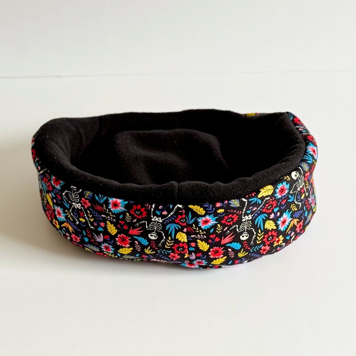 The Blooms & Bones Fleece Bed—a cozy, round nest for guinea pigs and small pets—features a velvety black fleece interior, perfect for midnight snuggles. Its exterior is draped in vivid floral patterns that bloom like enchanted wildflowers against the night. The bed is pictured resting on a bone-white surface, casting soft shadows worthy of gothic fairy tales.