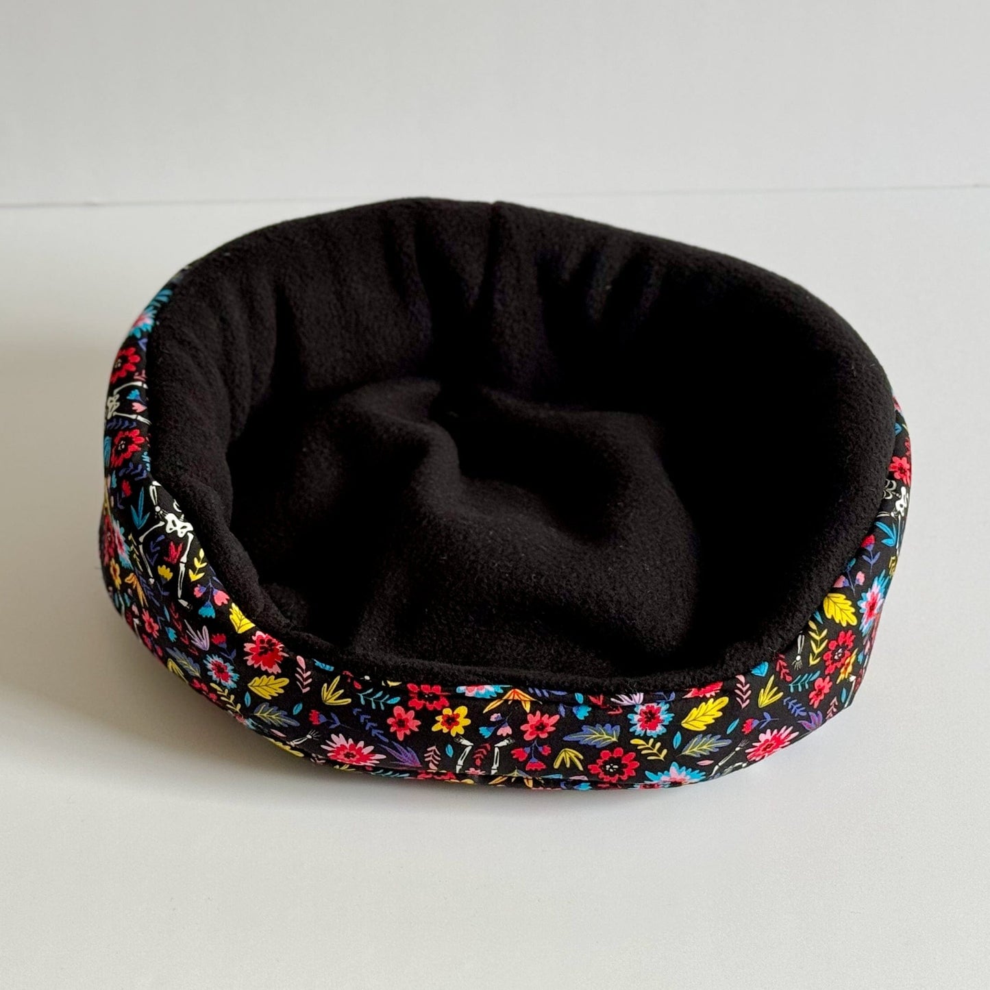 The Blooms & Bones Fleece Bed enchants with midnight-black plush inside and a vivid border of gothic florals in magenta, teal, and emerald, swirling around the cozy round shape. Perfectly sized for guinea pigs or small familiars to curl up in spooky-cute comfort. Displayed on an ethereal white backdrop, this bed whispers glam charm fit for your little creature of the night.