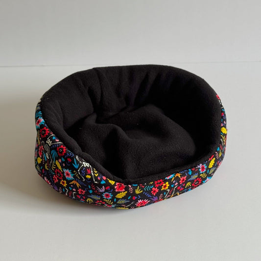 The Blooms & Bones Fleece Bed beckons from the shadows—a luxuriously plush, round nest in deep midnight black, crowned with a gothic-glam rim of enchanting florals and delicate skeletal motifs. This cozy haven invites your guinea pig or pampered small pet to lounge in moonlit comfort and mysterious style, all atop crisp white surfaces.