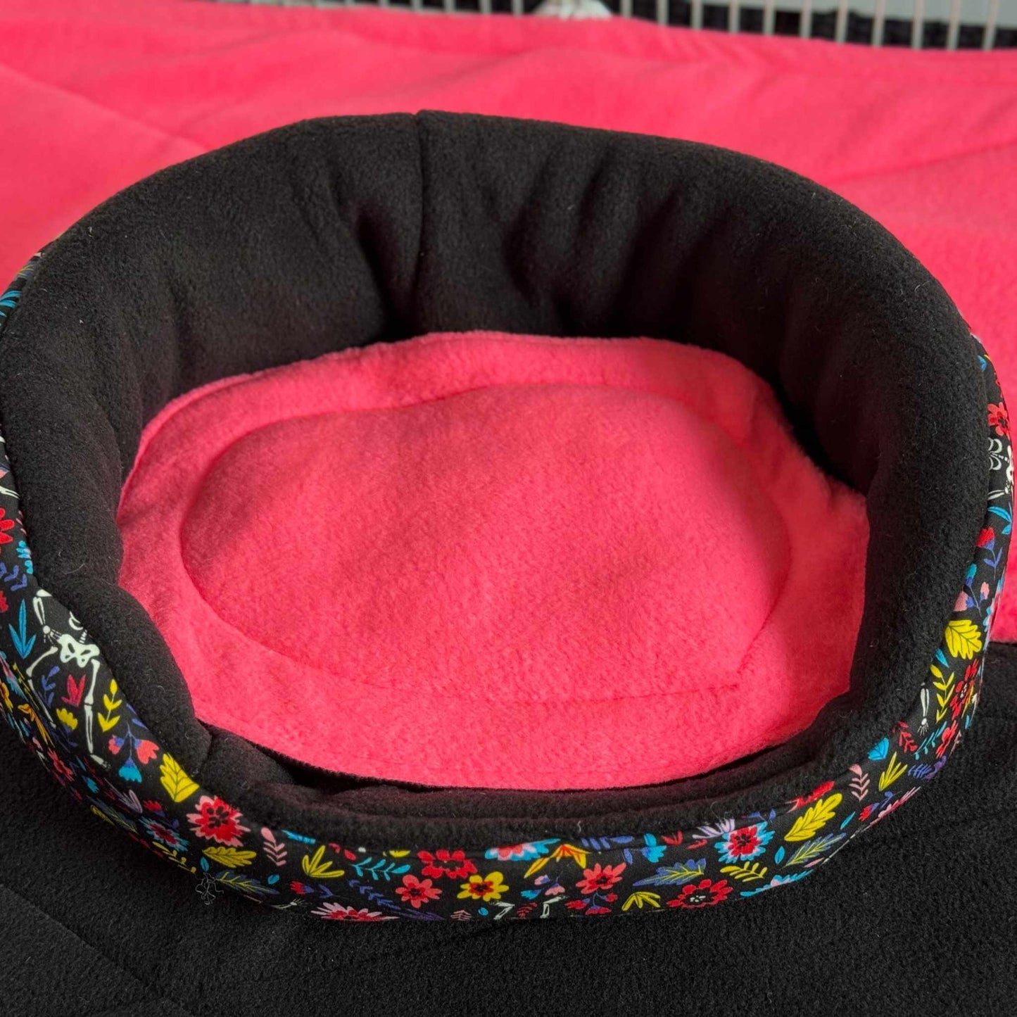 Enchant your guinea pig with a gothic-glam Blooms & Bones Bed—round, bright pink cushion, eerie floral-black sides, and cozy fleece blanket.