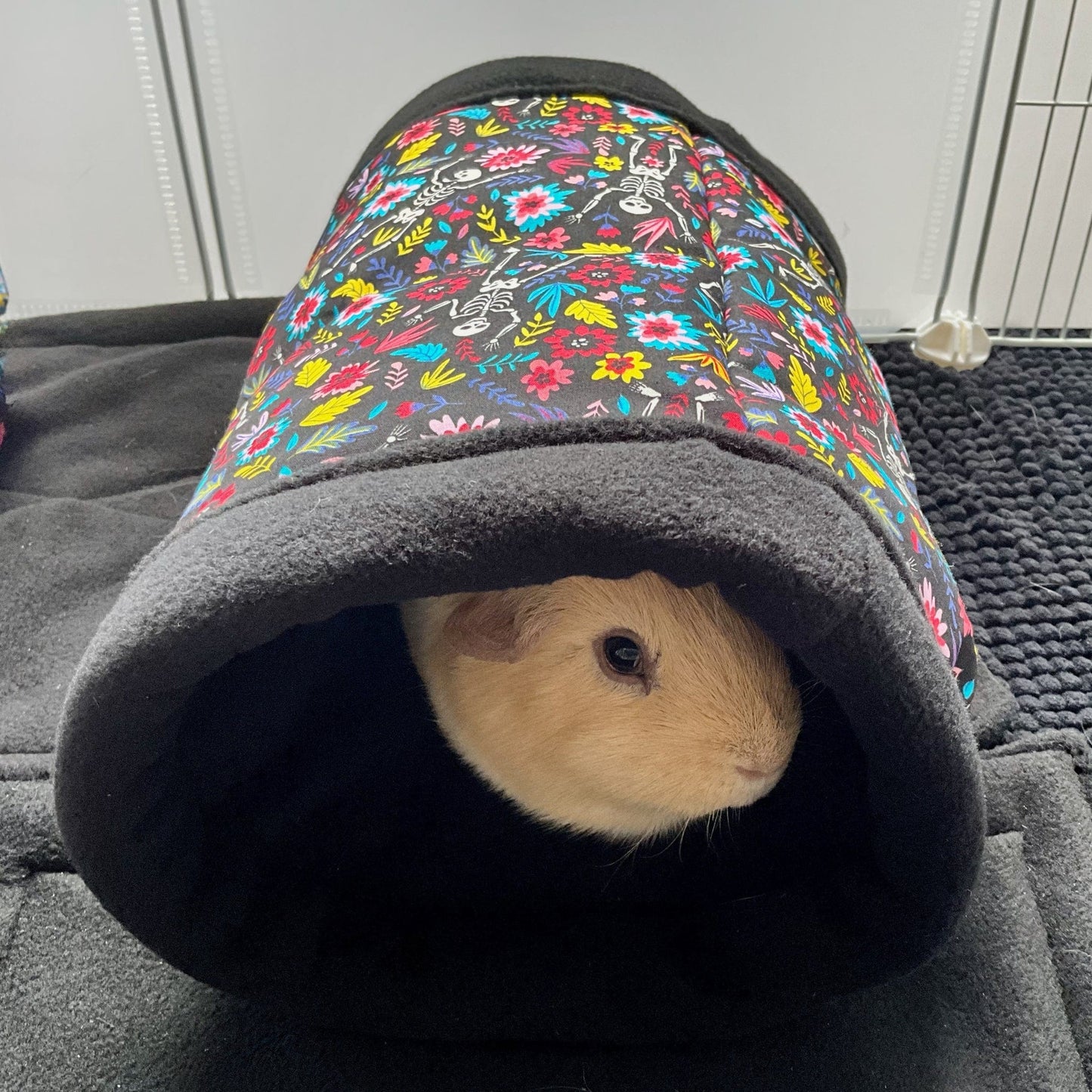 A sweet, cinnamon-hued guinea pig peeks enchantingly from within the Blooms & Bones Fleece Tunnel—a plush, stay-open burrow cloaked in a gothic tapestry of vivid blossoms entwined with whimsical skeletons. The little explorer lounges atop a shadowy mat, surrounded by florals and bones that whisper dark fairytales, creating the perfect haunt for your charmingly mysterious companion.