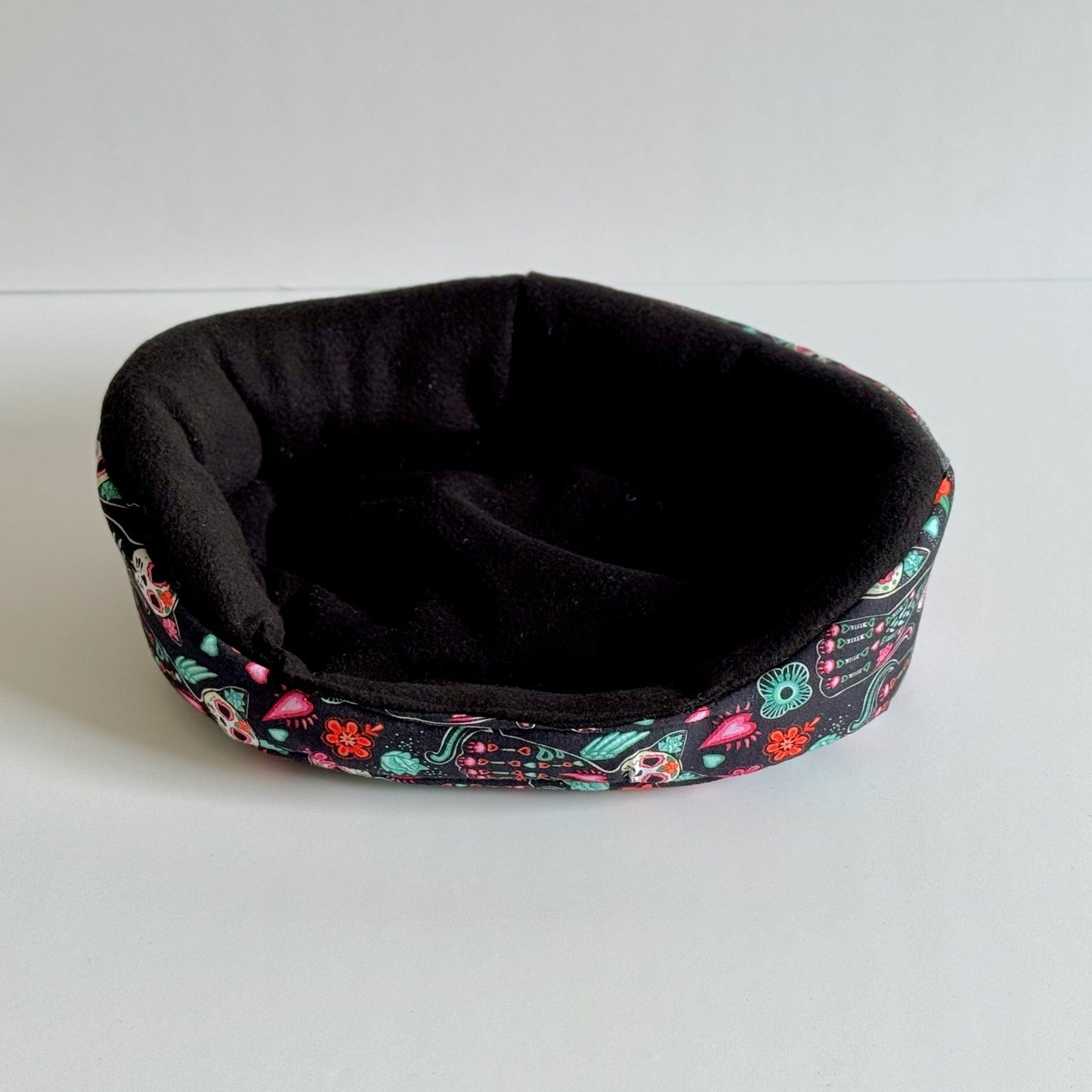 The Curious Catrina Fleece Bed enchants with a midnight-black, ultra-plush nest inside, while the outside dazzles in gothic glam—swirls of vivid flowers and whimsical sugar skulls dancing across deep fabric. This cozy, small round bed beckons adventurous guinea pigs and tiny familiars to curl up in spooky-cute comfort. Displayed on a simple white background for a hauntingly charming contrast.