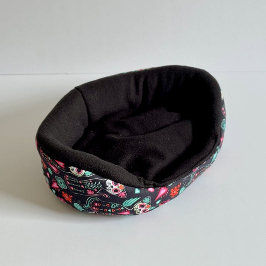 The Curious Catrina Fleece Bed—a whimsically gothic nest for your beloved small pet—features a plush, midnight-black interior perfect for curling up in secretive comfort. The exterior is bedecked in an enchanting tapestry of vibrant sugar skulls, blooming flowers, and twisting leaves on a dark canvas, conjuring Dia de los Muertos magic with every stitch. This oval-shaped haven is equal parts cozy crypt and glam hideaway, displayed here on an ethereal white background.