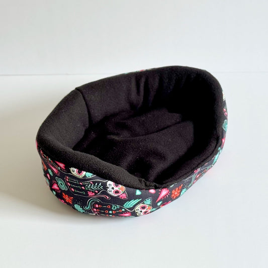 Enchant your tiny familiar with a cozy black snuggle sack shrouded in vivid skulls and blooms—spooky-glam comfort awaits!.