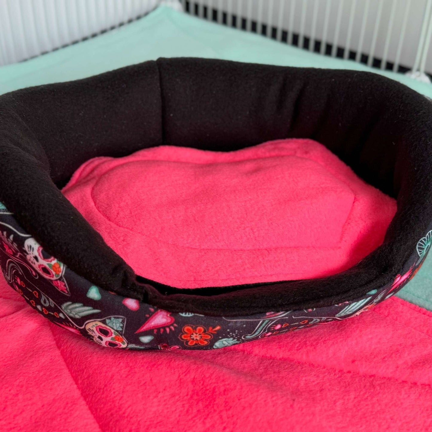 The Curious Catrina Fleece Bed beckons your beloved guinea pig into a world of spooky-cute luxury—nestled atop a bold turquoise stage, its plush bubblegum-pink interior contrasts dramatically with a black rim adorned in whimsical skulls and moonlit florals. This gothic-glam snuggle spot weaves cozy comfort with mysterious charm, ready to enchant tiny paws. Shown resting near a softly glowing white wall.