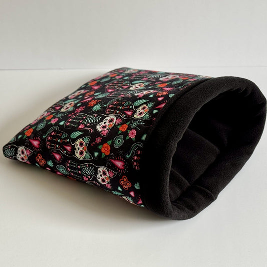 The Curious Catrina Snuggle Sack—a plush, midnight-black fleece tunnel—invites your guinea pig to slumber in gothic-glam style. Its outer fabric dazzles with whimsical Day of the Dead sugar skulls in vibrant hues, twined with enchanting floral patterns. Perfect for cozy naps, this spooky-cute hideaway is pictured resting atop a snowy white background, ready to cradle your small pet in mysterious comfort.