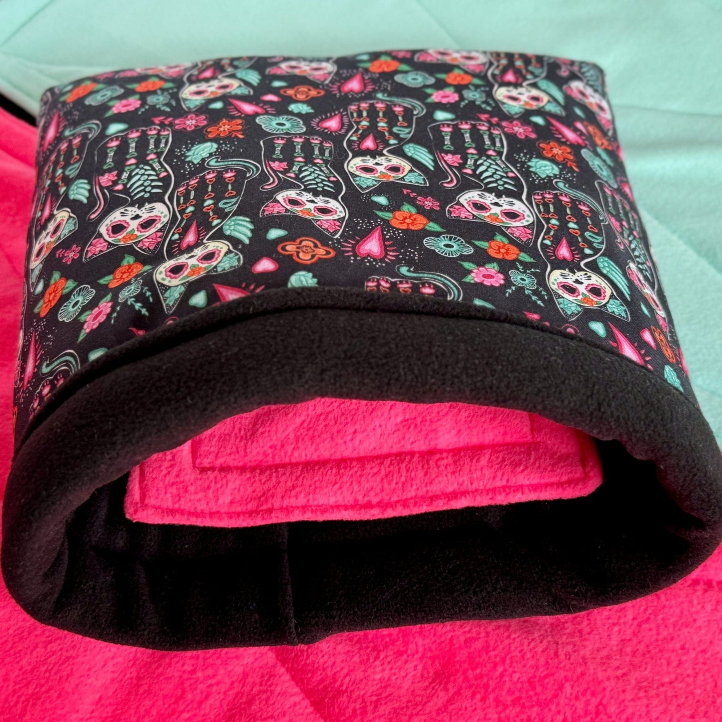 The Curious Catrina Snuggle Sack—a cozy hideaway for guinea pigs—features a midnight-black exterior cloaked in vibrant sugar skull kitties, their eyes sparkling with gothic charm. Peek inside to find plush, vivid pink fleece for a pop of playful color. Pictured rolled witchily on a mint green stage, ready to cradle your little darling in spooky-cute luxury.