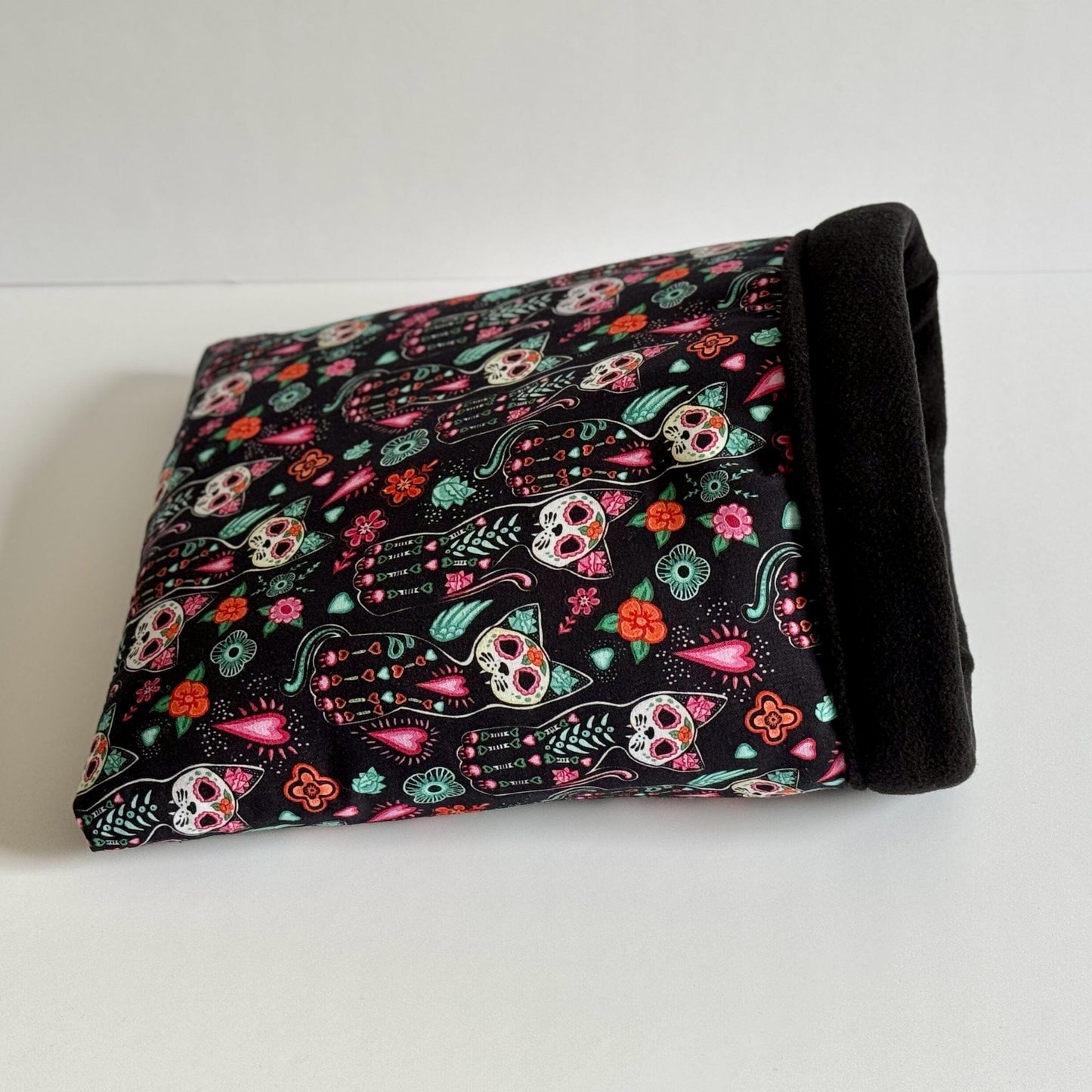 The Curious Catrina Snuggle Sack beckons with velvety black fleece, adorned in a lively parade of gothic-glam sugar skulls and hauntingly bright flowers—perfect for your small creature of the night. This pet hideaway, fit for any whimsical guinea pig or tiny familiar, lies invitingly folded atop a moon-pale white backdrop, ready to cradle spooky dreams and cozy snuggles.