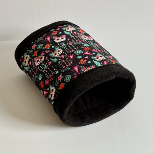 The Curious Catrina Fleece Tunnel—a gothic-glam hideaway for guinea pigs—bewitches with inky black fleece adorned in enchanting sugar skull cats, their faces blooming with whimsical flowers and charming heart motifs. Vibrant colors pop against the dark fabric, conjuring a delightful daydream of spooky sweetness. Tunnel rests invitingly on a crisp white backdrop, ready to host your little pet’s mysterious midnight (or midday) adventures.