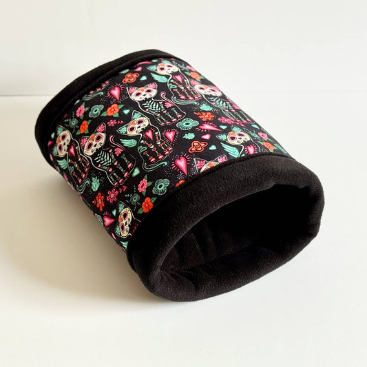 The Curious Catrina Fleece Tunnel—a bewitchingly cozy hideaway for small pets—bewitches with gothic glamour: jet-black fleece adorned with radiant Day of the Dead cat skulls, swirling in kaleidoscopic purples, hot pinks, and electric blues. Playful flowers bloom alongside the spooky-cute felines across soft, plush sides. This pint-sized tunnel is fully lined and machine washable, pictured here lounging atop a ghostly white backdrop—perfect for charming your tiny familiars.