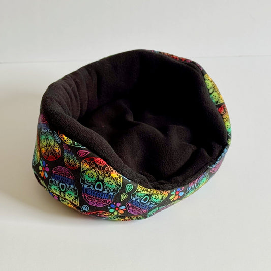 The Frightful Fiesta Fleece Bed beckons your small companion to drift into moonlit dreams—a cozy square nest lined with plush midnight-black fleece. The outer sides bloom with gothic-glam vibrance, crowned in swirling sugar skulls and enchanted florals bursting with purples, pinks, and haunting gold. Perfectly sized for guinea pigs or pocket-sized familiars seeking a whimsically spooky snooze spot. Shown resting on a stark white background for an extra dash of dramatic contrast.