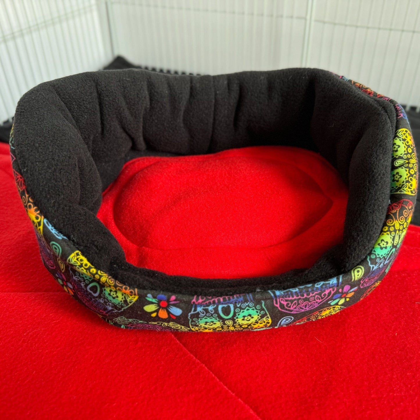 The Frightful Fiesta Fleece Bed—a gothic-glam haven for your little familiars—boasts a plush, midnight-black interior that whispers cozy secrets. Its exterior bursts with a whimsical carnival of jewel-toned patterns, casting a spell of vibrant color beneath pale moonlight. The bed lounges atop blood-red fleece inside a crisp white wire enclosure, creating the perfect haunted palace for guinea pigs or other pint-sized companions craving comfort and style.