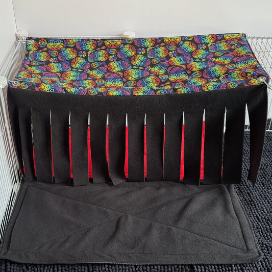 Inside a white wire castle, cozy shadows fall beneath a whimsical roof stitched with rainbow owls—wide-eyed guardians of the night. A Frightful Fiesta Large 2x1 Fleece Forest curtain drapes dramatically, its black and orange strips beckoning tiny adventurers through gothic arches. The floor is layered with a plush, storm-gray mat, perfect for midnight scurries and drowsy daydreams alike. Perfectly eerie-chic for your enchanting little companion!.