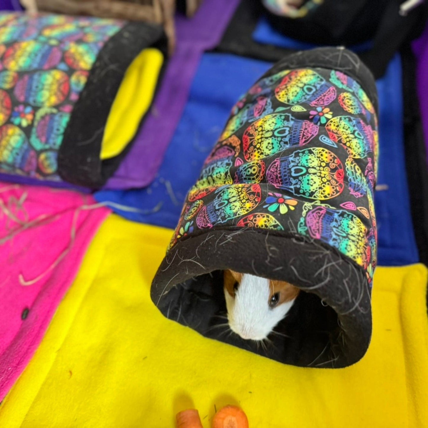 A curious guinea pig peeks out from a haunted carnival-patterned tunnel atop a golden-yellow mat. In front, a plump orange carrot awaits their nibble. Beside the scene, Frightful Fiesta Matching Pee Pads—cushy fleece pads hand-sewn with gothic glam in every stitch—promise enchanting, easy cleanups for your pampered beastie.