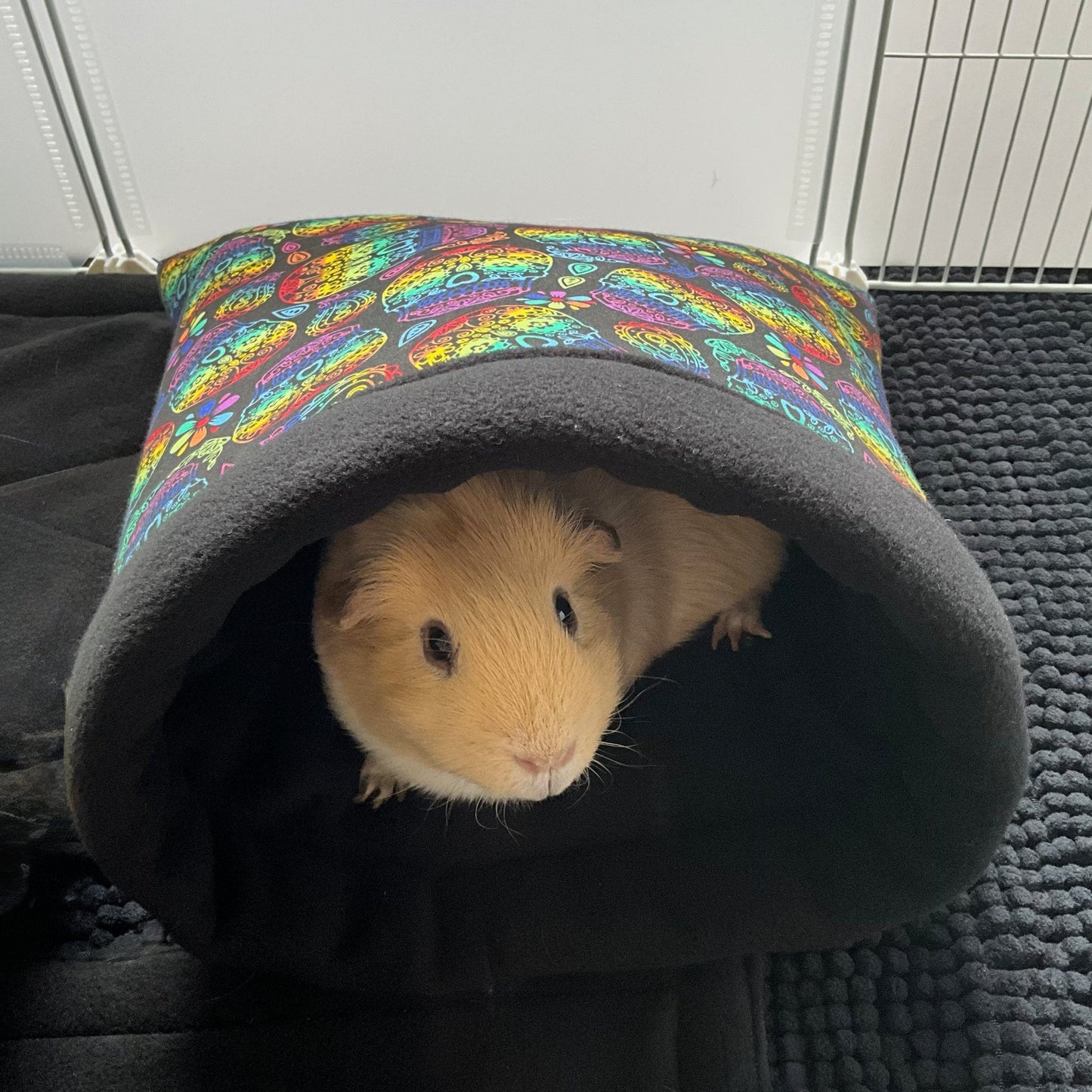 A caramel-hued guinea pig lounges like a gothic darling inside the Frightful Fiesta Snuggle Sack—its cozy cocoon adorned with vibrant, wickedly whimsical patterns and lined with midnight-soft fleece. The plush sack rests atop a shadowy black surface, with the delicate bars of a white metal cage casting ghostly lines in the background.