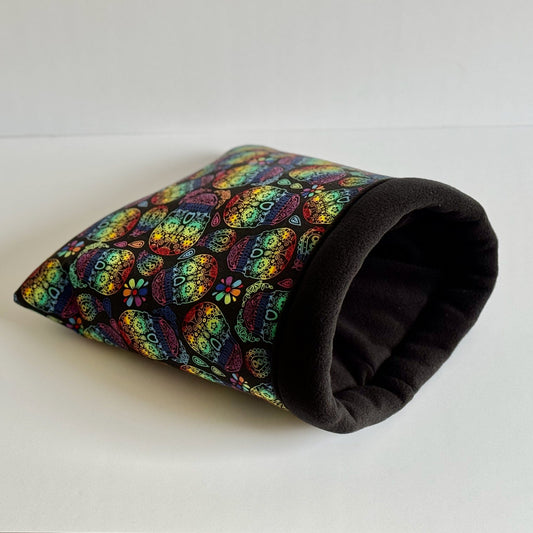 The Frightful Fiesta Snuggle Sack—an enchanting guinea pig bed bedecked in midnight-black, pumpkin orange, and ghostly violet patterns—beckons from a moonlit-white backdrop. Its plush fleece interior promises spine-tingling coziness for your small companion, offering a luxurious gothic-glam hideaway perfect for nocturnal snuggles and whimsical dreams.