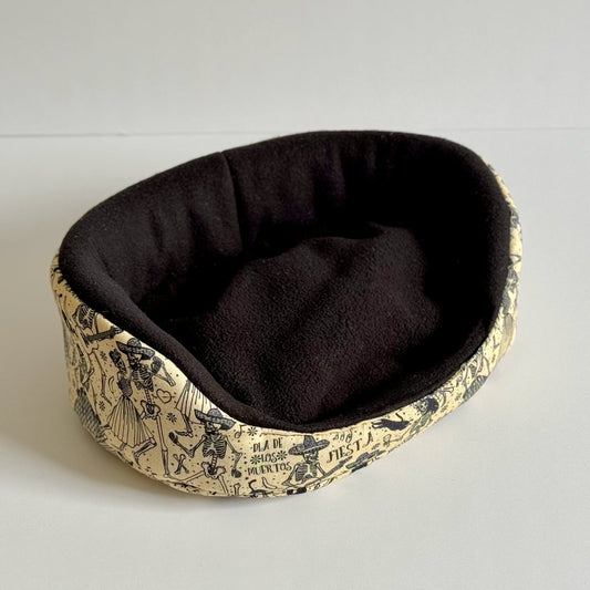 The Tombstone Tango Fleece Bed is a cozy, oval nest fit for your tiniest familiars—a plush black fleece cushion beckons from within, cradled by soft beige sides twirling with gothic tombstones, whimsical bats, and moonlit florals in inky black. Perfectly sized for guinea pigs or small animal ghouls, this bed summons spooky-cute charm to any lair. Shown resting atop a pale backdrop, ready to enchant your beloved beasties.