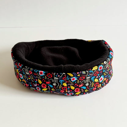 The Blooms & Bones Fleece Bed—a cozy, round nest for guinea pigs and small pets—features a velvety black fleece interior, perfect for midnight snuggles. Its exterior is draped in vivid floral patterns that bloom like enchanted wildflowers against the night. The bed is pictured resting on a bone-white surface, casting soft shadows worthy of gothic fairy tales.
