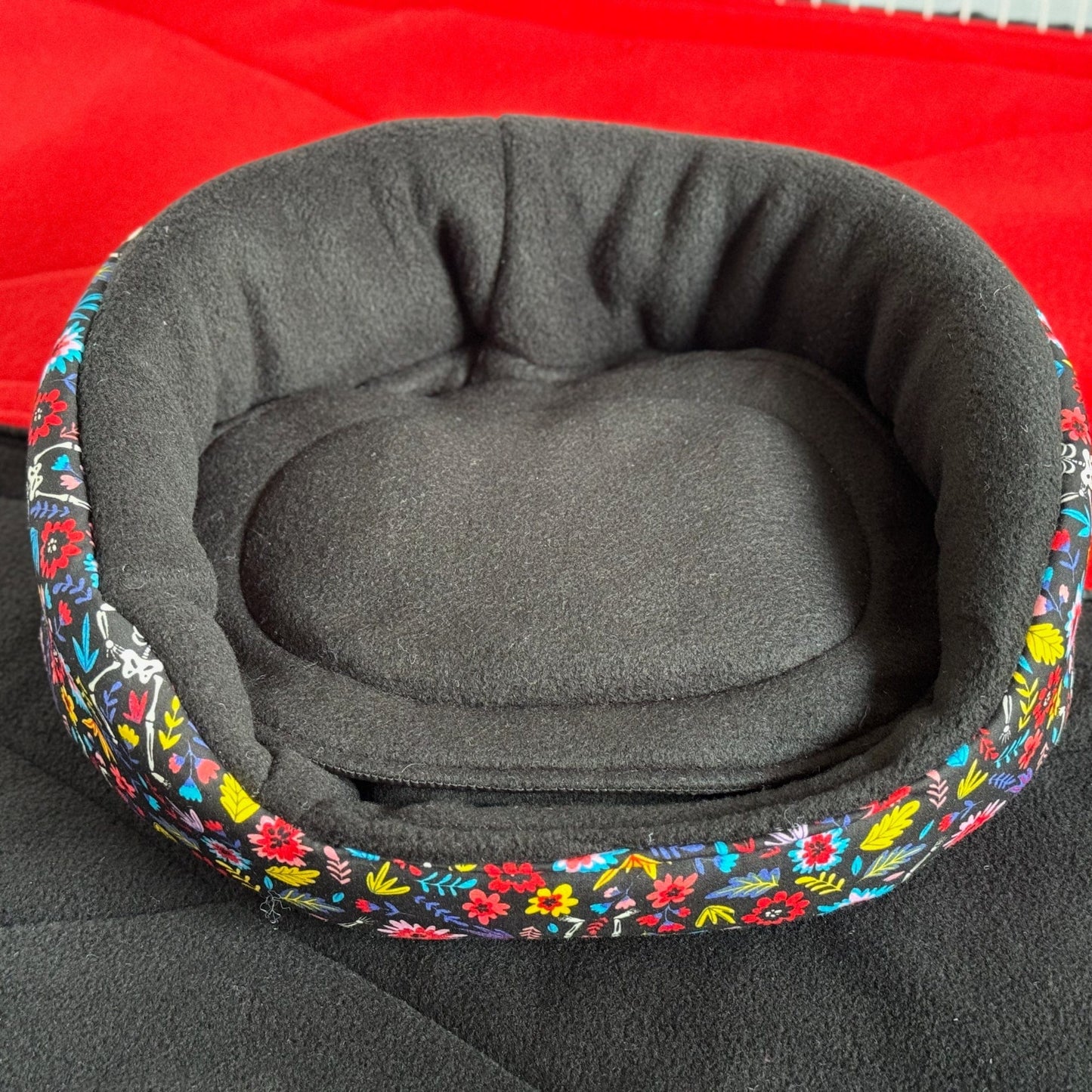 The Blooms & Bones Fleece Bed enchants with gothic glamour—an inky black, round nest perfect for your small familiar. Plush, high sides cradle tiny souls in cozy safety, while a lush, removable cushion invites the sweetest of crypt-side dreams. Adorned with a vibrant floral trim—a twining bouquet amid the midnight shadows—this bed brings moonlit mystique to any piggy palace. Shown here resting atop a crimson surface for an extra hint of haunted elegance.