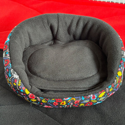 The Blooms & Bones Fleece Bed enchants with gothic glamour—an inky black, round nest perfect for your small familiar. Plush, high sides cradle tiny souls in cozy safety, while a lush, removable cushion invites the sweetest of crypt-side dreams. Adorned with a vibrant floral trim—a twining bouquet amid the midnight shadows—this bed brings moonlit mystique to any piggy palace. Shown here resting atop a crimson surface for an extra hint of haunted elegance.