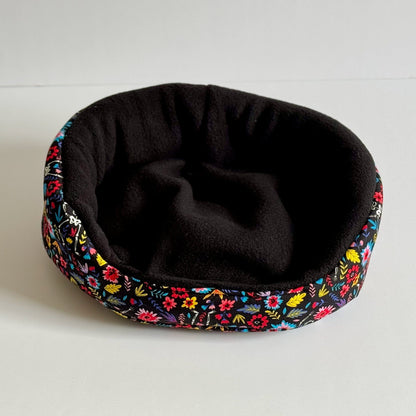The Blooms & Bones Fleece Bed enchants with midnight-black plush inside and a vivid border of gothic florals in magenta, teal, and emerald, swirling around the cozy round shape. Perfectly sized for guinea pigs or small familiars to curl up in spooky-cute comfort. Displayed on an ethereal white backdrop, this bed whispers glam charm fit for your little creature of the night.