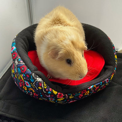 A sweet, light brown guinea pig perches upon our Blooms & Bones Fleece Bed—a gothic-glam nest woven from midnight-black fleece adorned with enchanting, jewel-toned flowers and dainty white bones. Nestled atop a plush scarlet cushion, this little companion gazes off to the right, wrapped in cozy luxury fit for pet royalty.