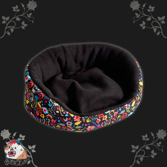The Blooms & Bones Fleece Bed is a bewitchingly cozy, round plush bed in inky black, ringed with whimsically bright blooms—floral patterns swirling along the edges like a garden under moonlight. Perfectly sized for guinea pigs or other small familiars, it invites naps with gothic glamour. Photographed atop a moody backdrop scattered with velvet flowers and charming cartoon critters lurking playfully in the shadows of each corner.