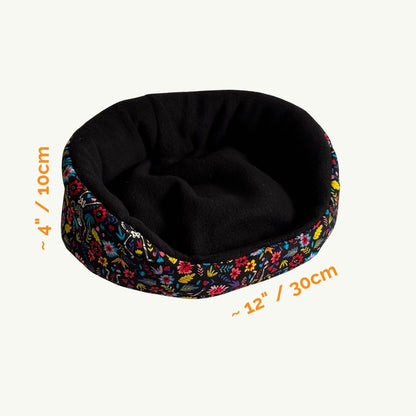 The Blooms & Bones Fleece Bed—a whimsically gothic nest for your beloved small pet! This cozy, round bed is shrouded in soft black fleece, crowned with a ruffled rim of enchanting florals in pinks, purples, greens, and blues—like a gothic garden at midnight. Sizing details glow in spooktacular orange script: approximately 4 inches (10 cm) tall and 12 inches (30 cm) wide. Perfect for guinea pigs seeking both comfort and hauntingly cute style!.