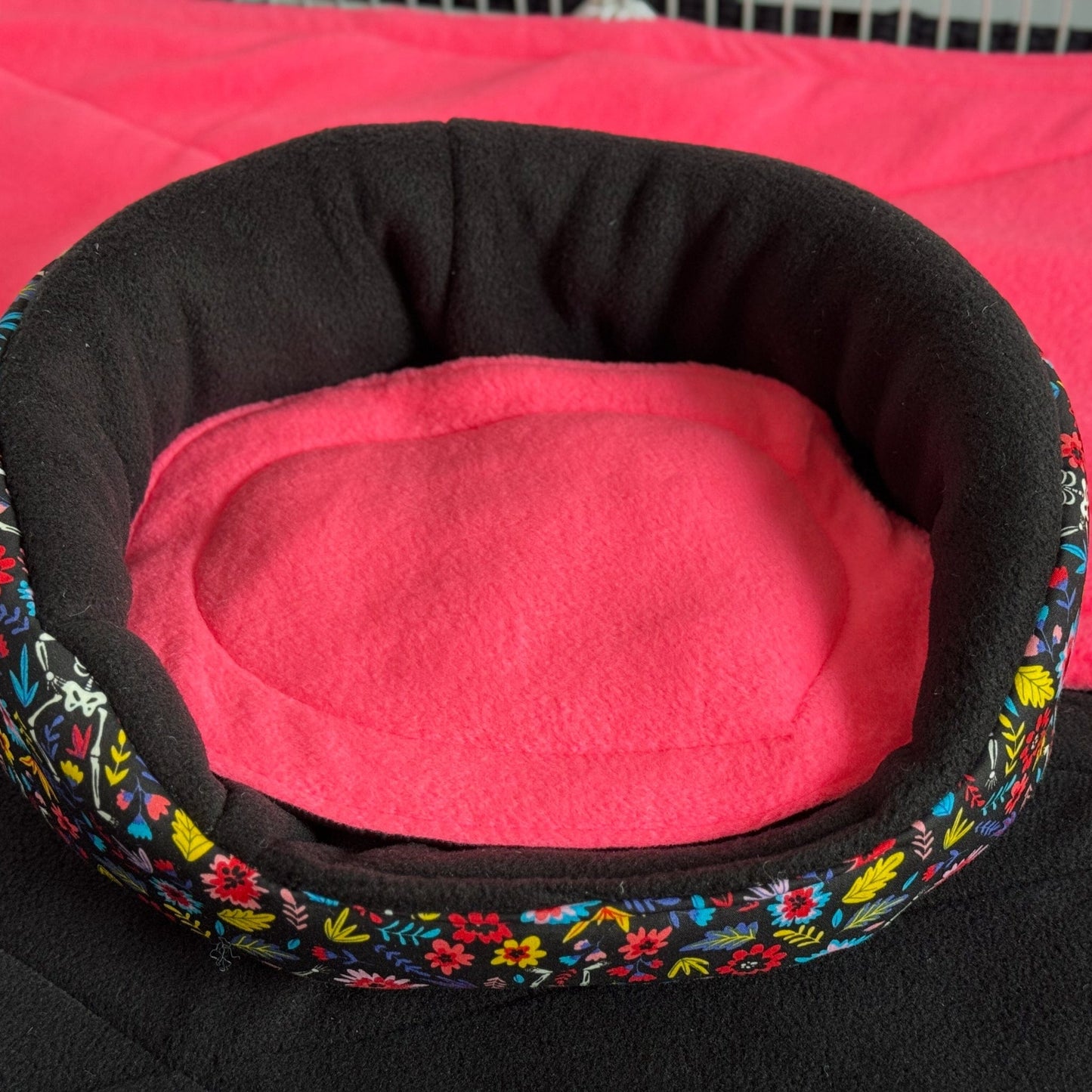 The Blooms & Bones Fleece Bed—a bewitching round nest for guinea pigs—features an enchanting black floral trim swirling with gothic elegance, cradling a plush, vivid pink fleece center. This cozy haven rests atop a matching pink-and-black backdrop, conjuring visions of midnight gardens and candy-colored dreams for your small pet’s slumber.