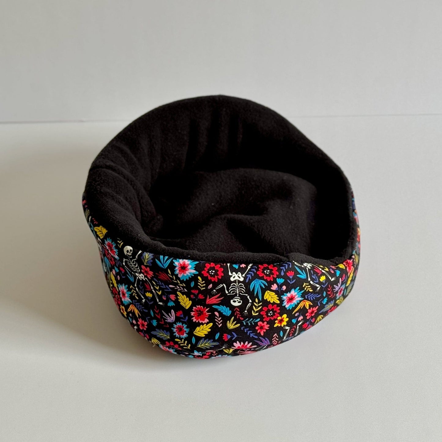 The Blooms & Bones Fleece Bed whispers gothic charm—a plush, round bed cloaked in inky black fleece. Its rim enchants with vibrant blossoms swirling around dainty skeletons, intertwining florals and bone motifs in a whimsigoth celebration. Perfectly sized for your fancy guinea pig or petite familiar. Pictured on a moon-pale white background for hauntingly chic contrast.