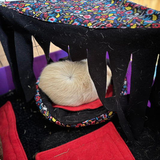 A creamy white guinea pig lounges, half-enchanted in shadow, beneath the Blooms & Bones Corner Fleece Forest—a whimsical woodland of midnight-black fleece adorned with gothic floral blooms. Petals and bones intertwine on fabric curtains that cascade from above, creating a mysterious yet cozy cavern in the cage’s corner. Blood-red fleece pads beckon like velvet rose petals on the floor nearby, perfect for tiny paws to rest upon.