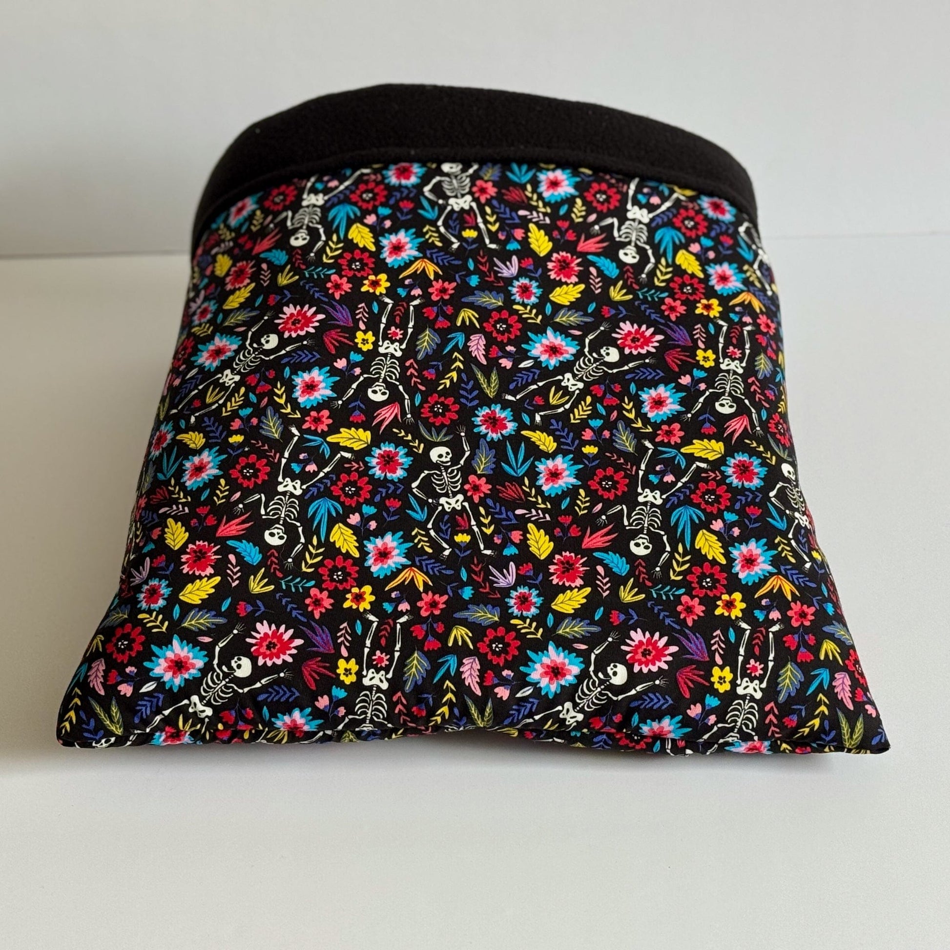 The Blooms & Bones Snuggle Sack enchants with a cryptic-chic flair—its rectangular shape is draped in midnight-black fabric, bedecked with vivid gothic blooms and mischievous little skeletons dancing among the petals. The top edge boasts an elegant band of deep black, while inside awaits a plush, jet-black fleece lining to cradle your guinea pig or small companion in cozy luxury. Shown resting atop a moon-pale white background for dramatic contrast.