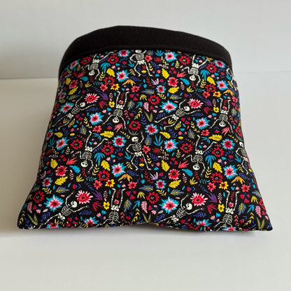 The Blooms & Bones Snuggle Sack enchants with a cryptic-chic flair—its rectangular shape is draped in midnight-black fabric, bedecked with vivid gothic blooms and mischievous little skeletons dancing among the petals. The top edge boasts an elegant band of deep black, while inside awaits a plush, jet-black fleece lining to cradle your guinea pig or small companion in cozy luxury. Shown resting atop a moon-pale white background for dramatic contrast.