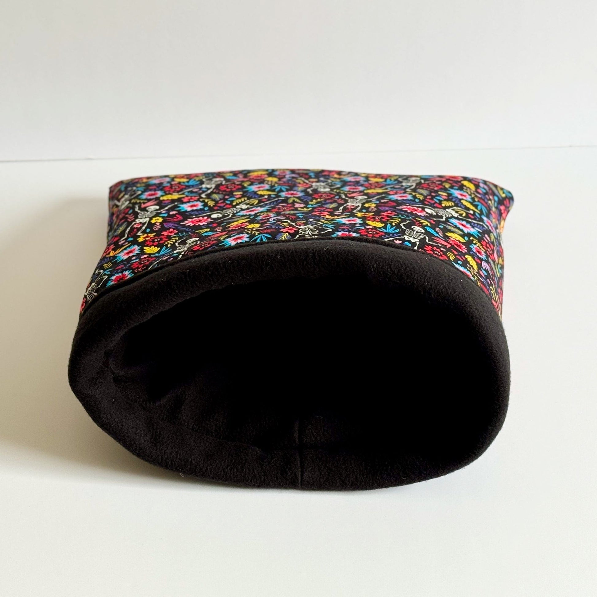The Blooms & Bones Snuggle Sack—a cozy, gothic-glam haven for guinea pigs and small pets—lies temptingly on its side atop a moonlit white surface. Midnight-black fleece wraps the snuggle sack, bedecked with enchanting pops of colorful gothic florals and whisperings of whimsy. Perfect for tiny familiars seeking a soft, mysterious hideaway beneath candlelight shadows.