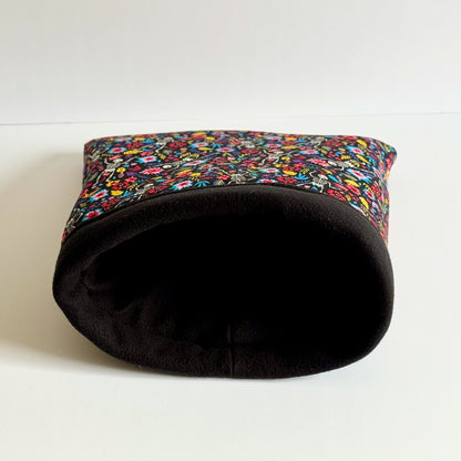 The Blooms & Bones Snuggle Sack—a cozy, gothic-glam haven for guinea pigs and small pets—lies temptingly on its side atop a moonlit white surface. Midnight-black fleece wraps the snuggle sack, bedecked with enchanting pops of colorful gothic florals and whisperings of whimsy. Perfect for tiny familiars seeking a soft, mysterious hideaway beneath candlelight shadows.