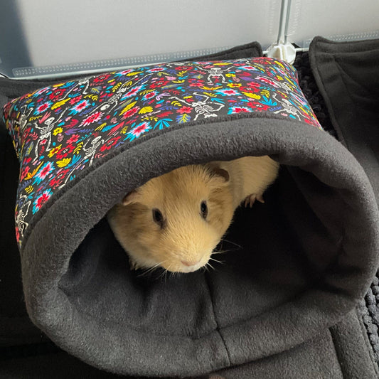 A light brown guinea pig gazes out from a Blooms & Bones Snuggle Sack, cozily ensconced in plush, midnight-black fleece. Vibrant gothic florals twine across the sack’s exterior—roses and moonlit leaves blooming upon a dusky backdrop—while stitched bone motifs add a touch of spooky whimsy. Wrapped in comforting enchantment, the little companion nestles safely inside this dreamy gothic-glam haven.