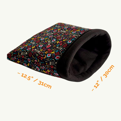The Blooms & Bones Snuggle Sack—a bewitching blend of midnight florals and mystical bone motifs—offers your darling guinea pig or small pet a luxuriously soft fleece hideaway. This gothic-glam tunnel, draped in vibrant blossoms upon an enchanting dark background, measures approximately 12.5 inches by 12 inches. Perfect as a charmingly eerie cuddle nook, shown invitingly against a pale, moonlit backdrop.