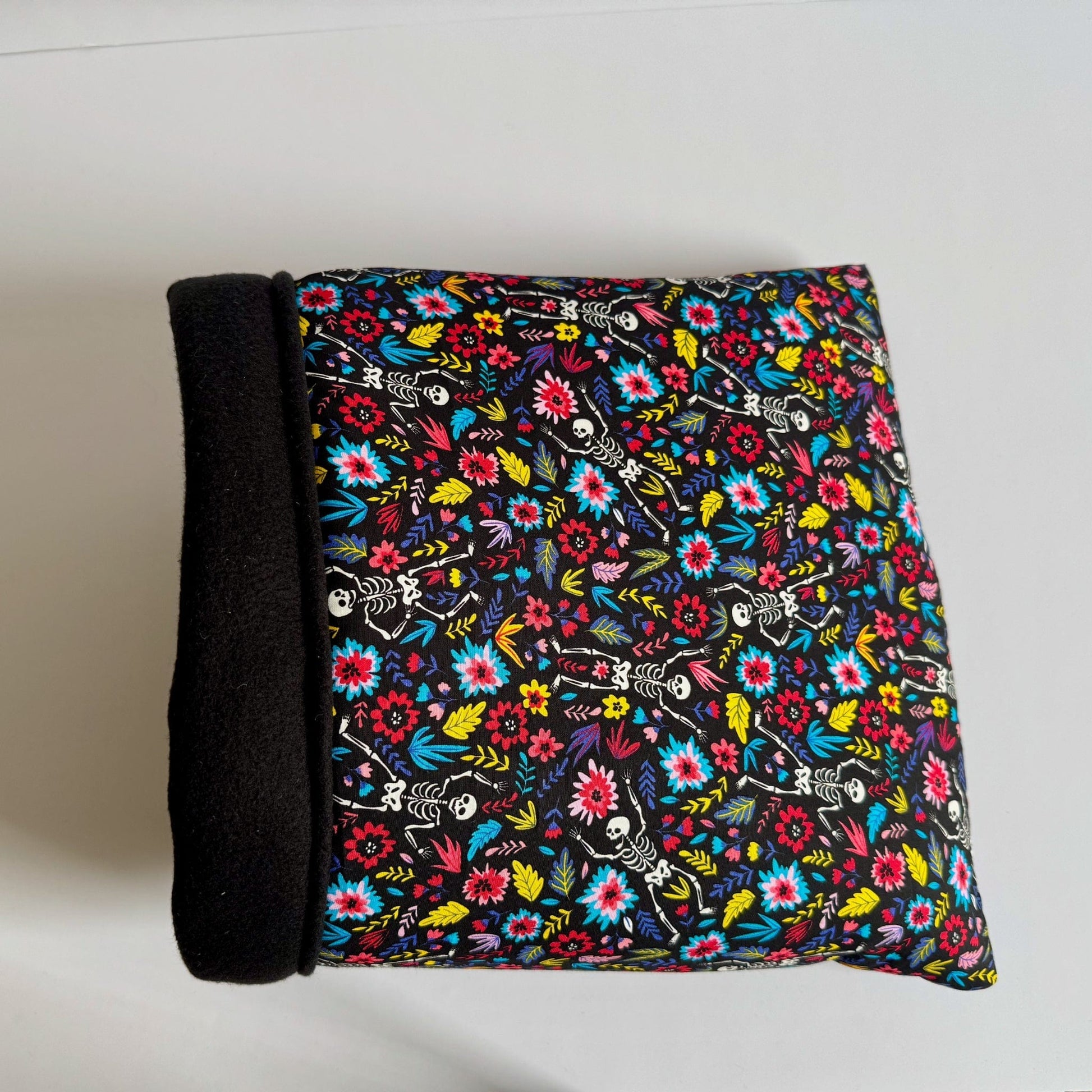 The Blooms & Bones Snuggle Sack—a plush rectangular hideaway for pets—features one bewitching black side and another draped in vivid florals intertwined with whimsical skeletons, evoking gothic garden dreams. Its interior nestles pets in midnight-soft fleece. Sack is pictured atop a ghostly white background.