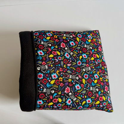 The Blooms & Bones Snuggle Sack—a plush rectangular hideaway for pets—features one bewitching black side and another draped in vivid florals intertwined with whimsical skeletons, evoking gothic garden dreams. Its interior nestles pets in midnight-soft fleece. Sack is pictured atop a ghostly white background.