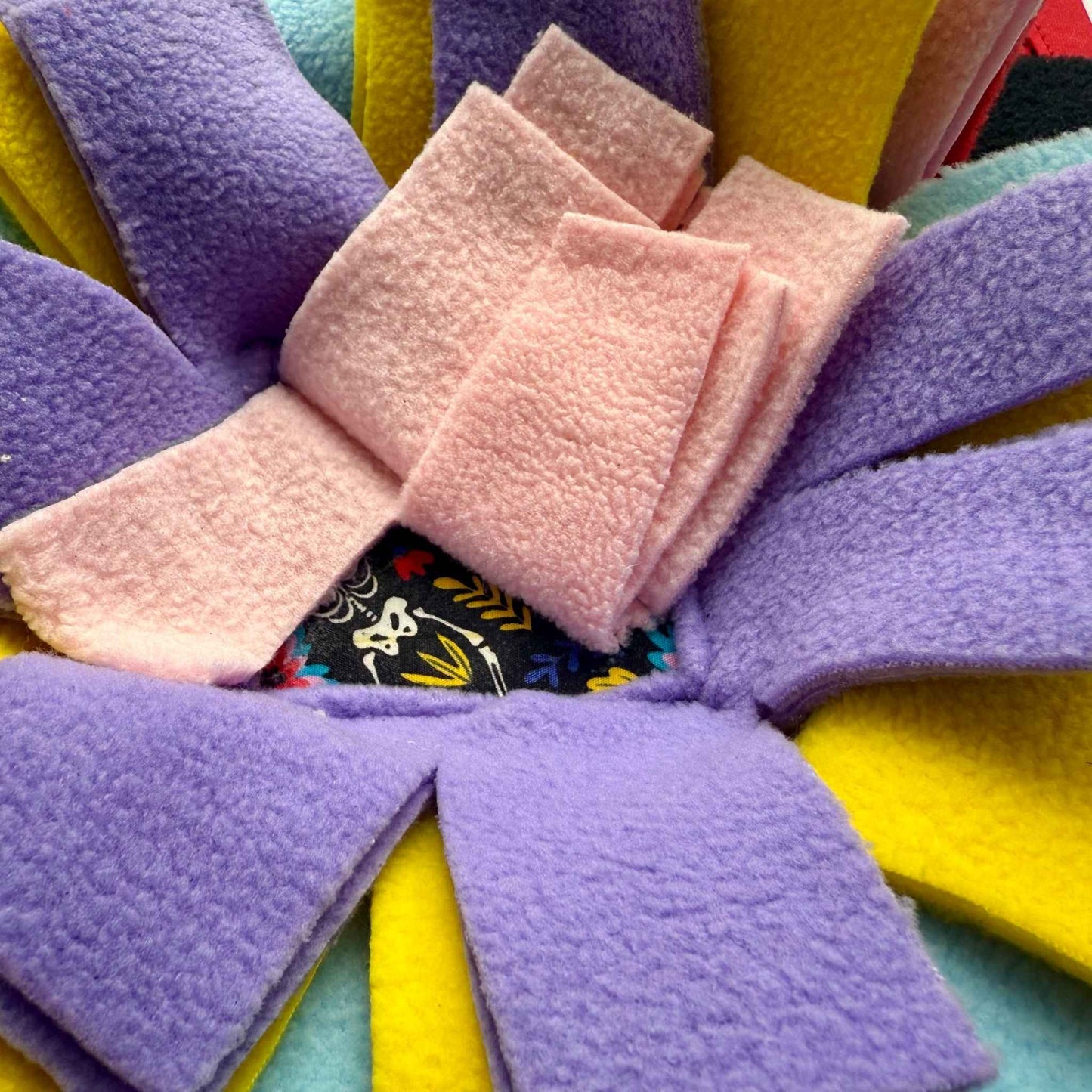 Blooms & Bones Snuffle Mat—vivid petals of yellow, purple, blue, and pink crowned by a pink bow on eerie patterned center.