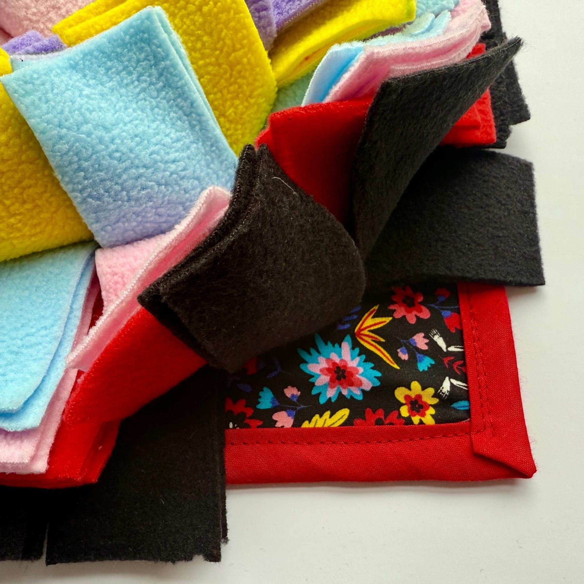 Gothic-glam snuffle mat close-up: candy-pink, midnight black, sapphire, and gold fleece atop a vivid floral print, ringed in crimson—spooky-cute for treat hunts!.