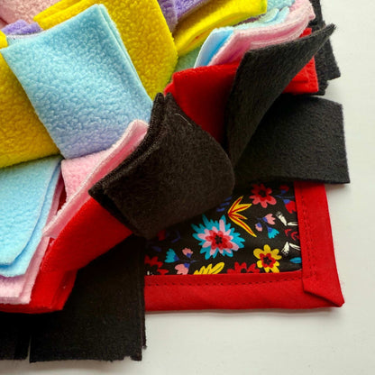 Gothic-glam snuffle mat close-up: candy-pink, midnight black, sapphire, and gold fleece atop a vivid floral print, ringed in crimson—spooky-cute for treat hunts!.