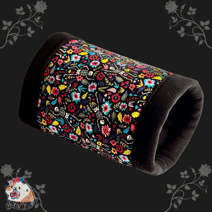 The Blooms & Bones Fleece Tunnel is a cozy, cylindrical hideaway for guinea pigs and small pets, bedecked in gothic-glam style. Black trim edges midnight-black fleece adorned with whimsical white skeletons entwined with moody floral vines. Set against a shadowy background bordered by dark, enchanted blossoms, a charming cartoon guinea pig peeks out from the corner—ready for spooky-cute snuggles in this mysterious little lair.