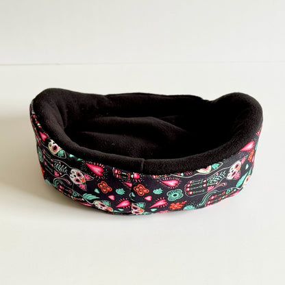 The Curious Catrina Fleece Bed beckons your little familiar to snuggle in gothic splendor—a plush black nest adorned with enchanting Day of the Dead sugar skulls and riotous, jewel-toned blooms. Whimsical skeleton faces wink among midnight petals, casting a spell of cozy comfort perfect for guinea pigs and other petite companions. Shown resting atop a moonlit white backdrop, this bed is equal parts spooky-cute haven and glam retreat for your small pets sweetest dreams.