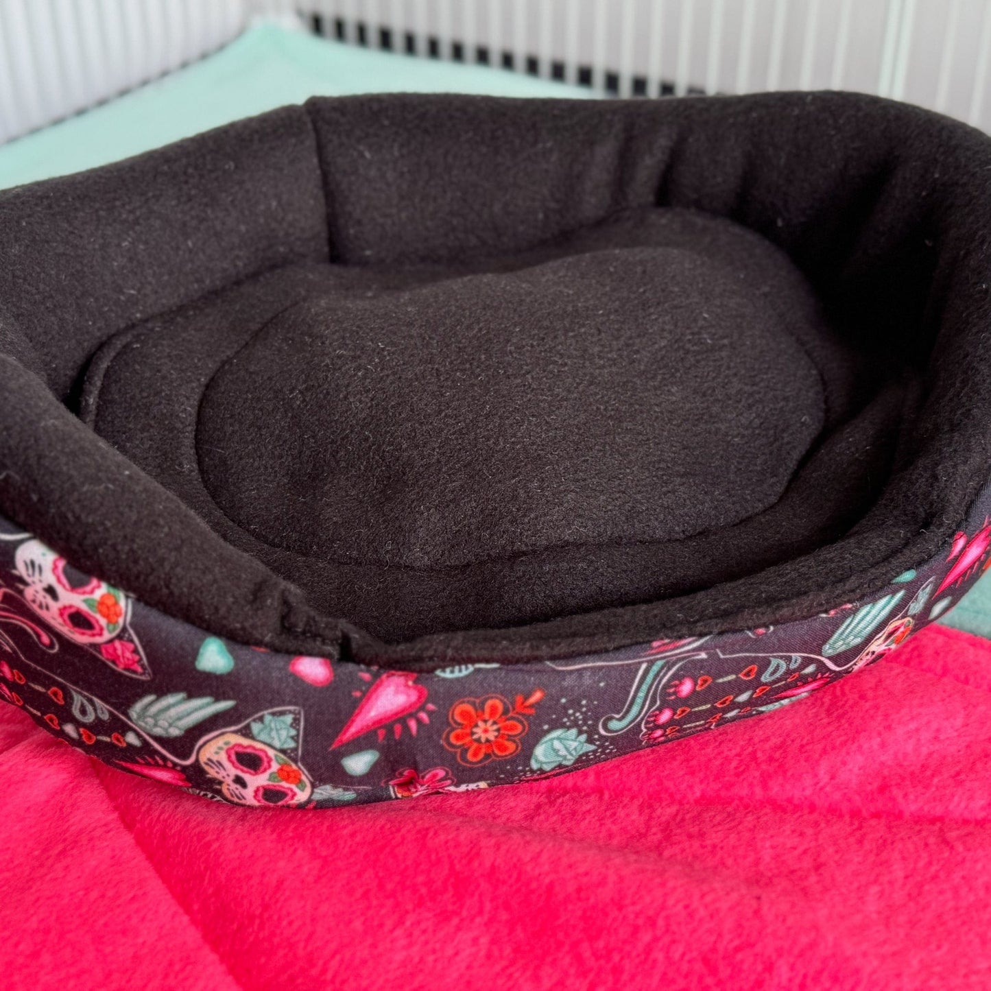 The Curious Catrina Fleece Bed awaits—a delightfully dark, black snuggle spot adorned with whimsical skulls and ornate gothic blooms. Perched atop a vivid pink blanket in a crisp white enclosure, this plush pet bed brings a splash of spooky-glamour for guinea pigs with mysterious charm and stylish dreams.