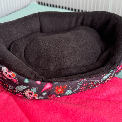 The Curious Catrina Fleece Bed awaits—a delightfully dark, black snuggle spot adorned with whimsical skulls and ornate gothic blooms. Perched atop a vivid pink blanket in a crisp white enclosure, this plush pet bed brings a splash of spooky-glamour for guinea pigs with mysterious charm and stylish dreams.