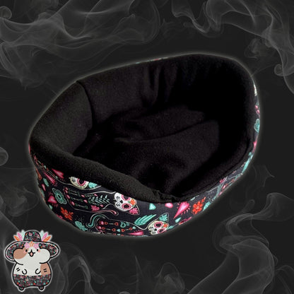 The Curious Catrina Fleece Bed enchants with its inky black oval shape, shrouded in a mysterious smoky backdrop. Bewitching sugar skull cats and vivid gothic florals twine around the sides—a lively little garden of daydreams for your small companion. In the lower left corner, a whimsical cartoon hamster tips its jaunty hat—ever the dapper greeter for guinea pigs or small critters daring to lounge in spook-tacular style!.