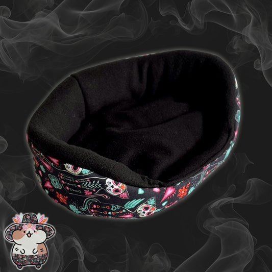 The Curious Catrina Fleece Bed enchants with its inky black oval shape, shrouded in a mysterious smoky backdrop. Bewitching sugar skull cats and vivid gothic florals twine around the sides—a lively little garden of daydreams for your small companion. In the lower left corner, a whimsical cartoon hamster tips its jaunty hat—ever the dapper greeter for guinea pigs or small critters daring to lounge in spook-tacular style!.