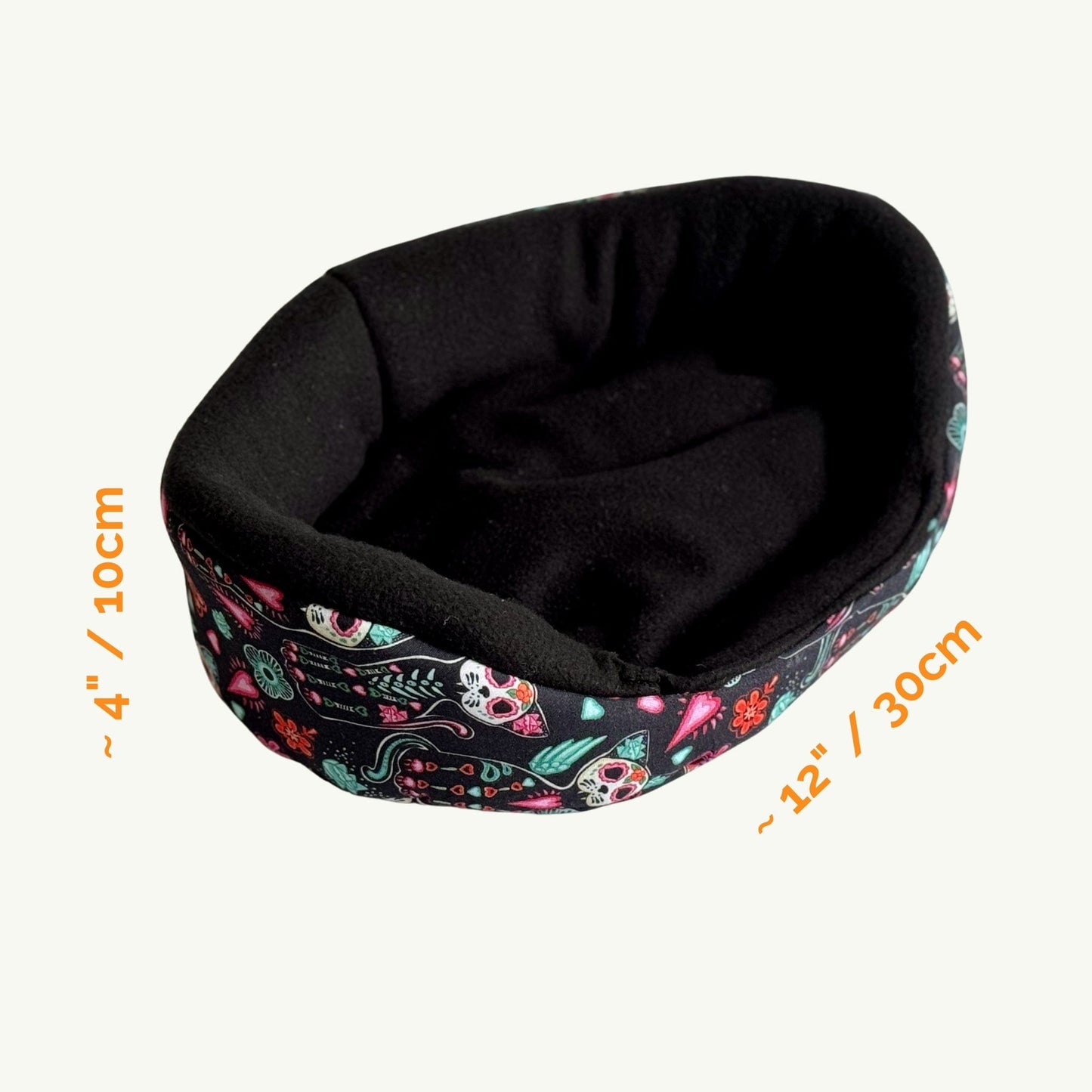 The Curious Catrina Fleece Bed, an enchanting oval nest cloaked in midnight-black fleece and bedecked with a vibrant, sugar-skull inspired print—think playful pops of purple, teal, and magenta roses swirling under moonlit bones. Sized for guinea pig royalty at about 4 inches (10 cm) tall and 12 inches (30 cm) long, this plush throne is pictured against a pale backdrop, just waiting to cradle your small companion in gothic-glam coziness.